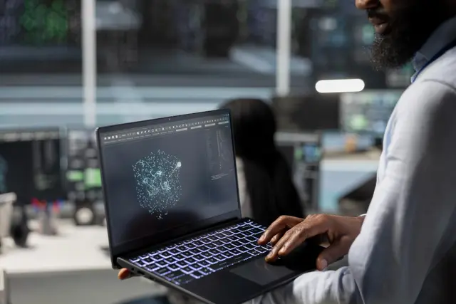 Bearded man holds an open laptop showing a network of glowing dots, in a room with screens in the background