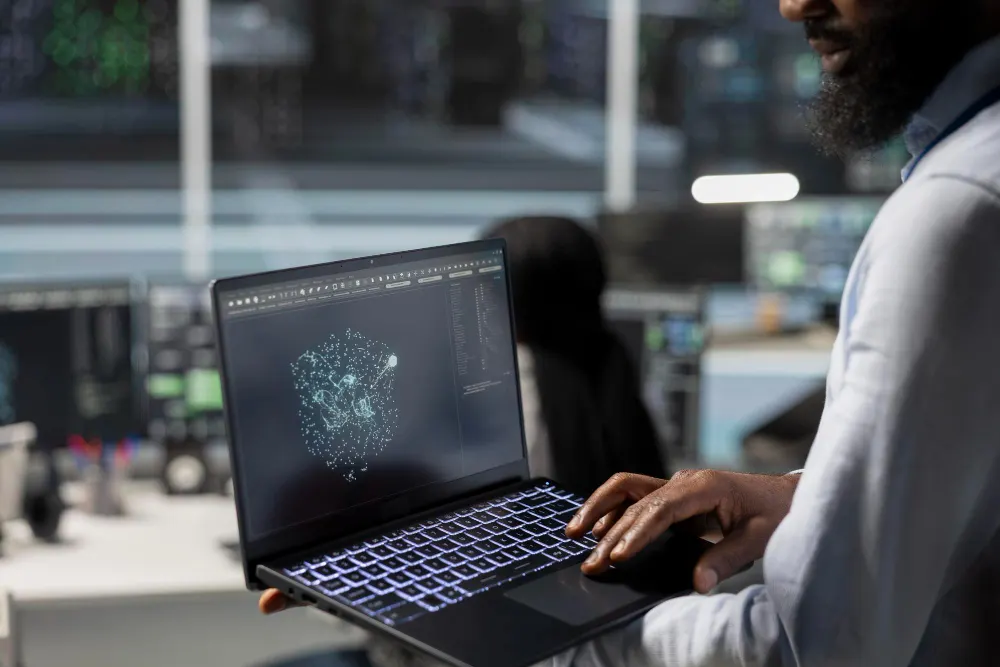 Bearded man holds an open laptop showing a network of glowing dots, in a room with screens in the background