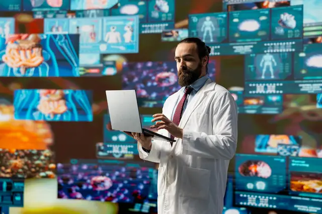 Doctor in a white coat and tie consults a laptop in front of a wall of screens showing anatomical and cellular images