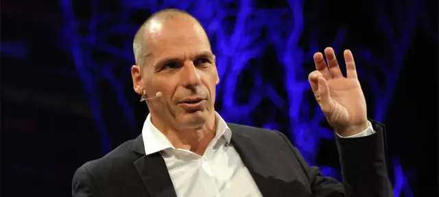 Yanis Varoufakis speaking on stage with a headset microphone and one hand raised, in front of a dark blue background with lights