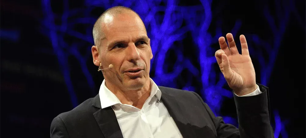 Yanis Varoufakis speaking on stage with a headset microphone and one hand raised, in front of a dark blue background with lights