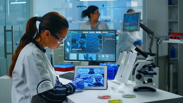 A scientist in a lab coat, gloves, and safety glasses looks at a tablet and a monitor showing blue microscopic images in a laboratory