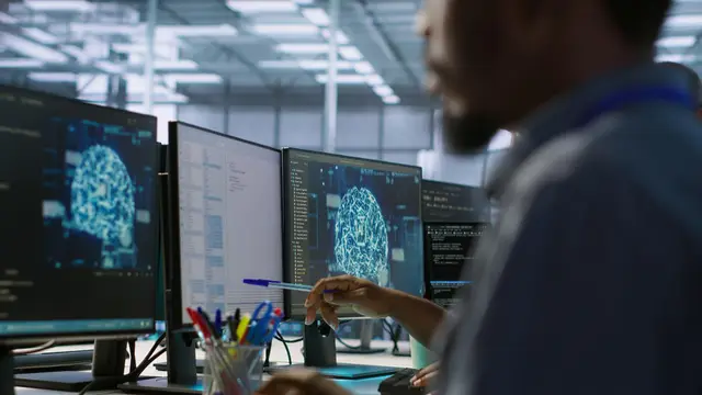 A man works in front of several monitors showing computer-generated images of a brain in a technology office