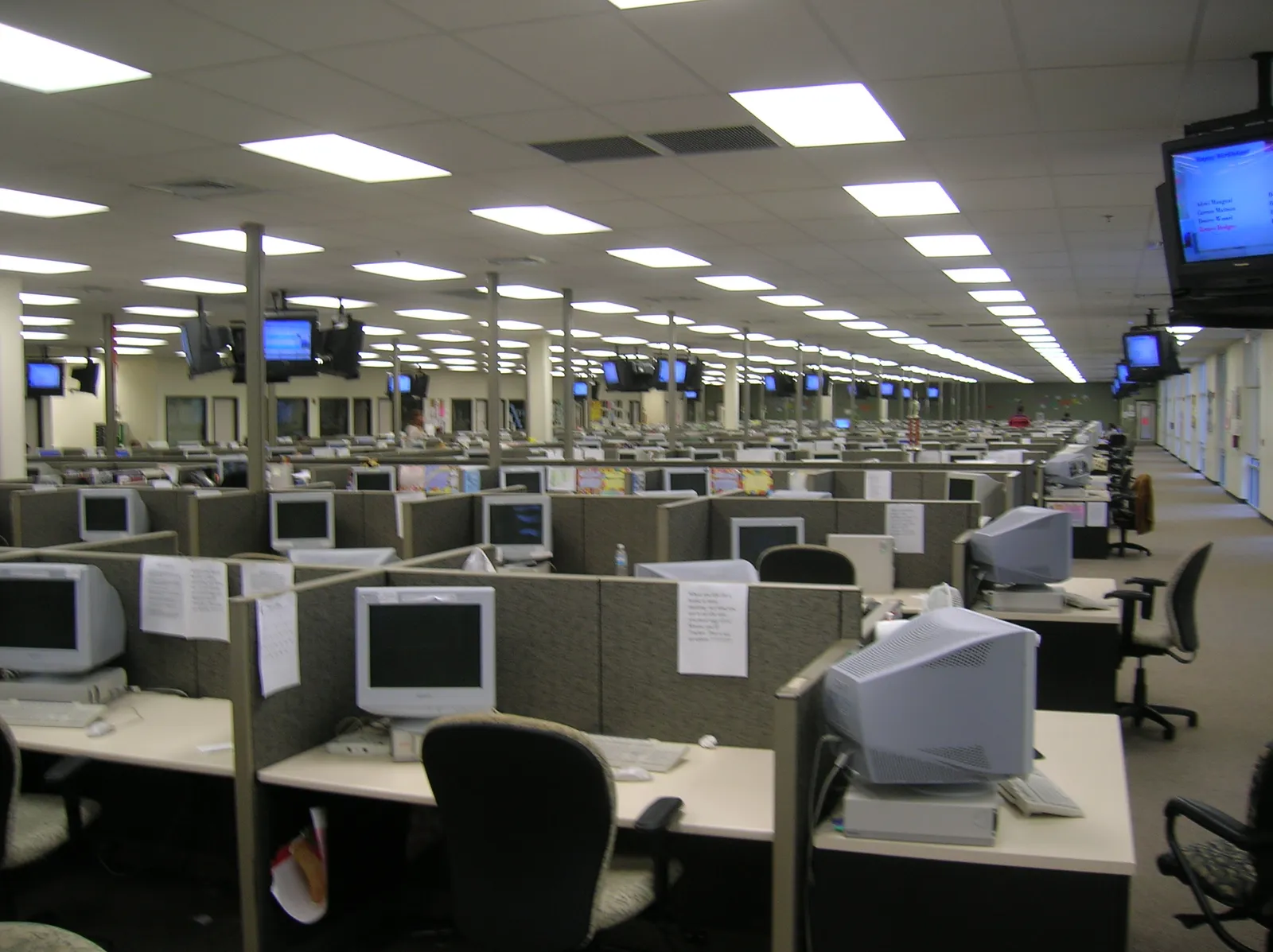 Rows of cubicles with CRT monitors, keyboards and chairs in a large office with rectangular ceiling lights.